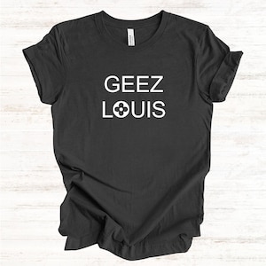 Könnte beinhalten: Ein dunkel anthrazitfarbenes Kurzarm-T-Shirt mit dem weißen Schriftzug GEEZ LOUIS auf der Brust, wobei ein vierblättriges Blumensymbol den Buchstaben O im Wort LOUIS ersetzt. Das T-Shirt hat einen Rundhalsausschnitt und eine Standardpassform.