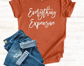Könnte beinhalten: Rostfarbenes T-Shirt mit dem weißen Schriftzug "Everything Expensive", kombiniert mit blauen Jeans. Die Jeans sind gefaltet und umgekrempelt, mit einem Distressed-Detail am Knie. Die Artikel sind auf einer hellen Holzmaserung angeordnet.