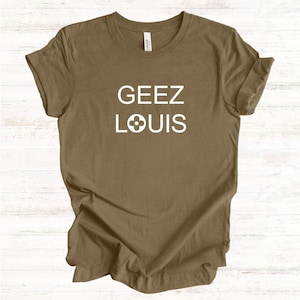 May include: A brown t-shirt with the words "GEEZ LOUIS" printed in white. The word "LOUIS" has a four-petal flower design in place of the "O". The shirt is made of a soft material.