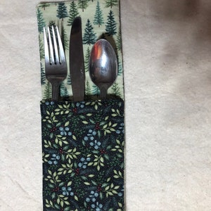 May include: A black fabric cutlery holder with a green and white floral pattern on one side and a green and white pine tree pattern on the other side. The holder contains a fork, knife, and spoon.
