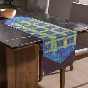 May include: A green, blue, and purple plaid table runner with a paisley design on the ends. The runner is on a black table with a wooden base.