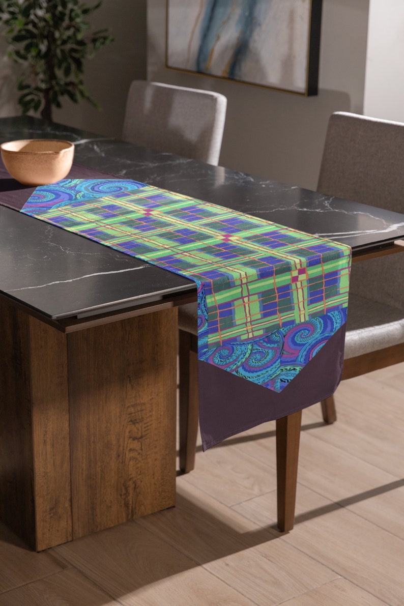 May include: A green, blue, and purple plaid table runner with a paisley design on the ends. The runner is on a black table with a wooden base.