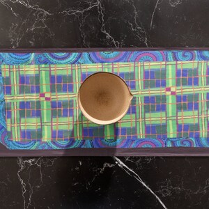 May include: A blue and green plaid table runner with a paisley border. A brown ceramic bowl is centered on the runner.