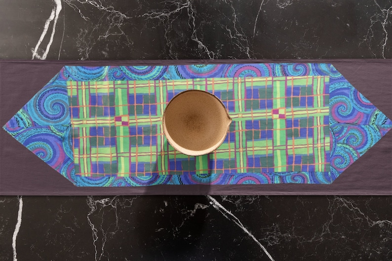 May include: A blue and green plaid table runner with a paisley border. A brown ceramic bowl is centered on the runner.