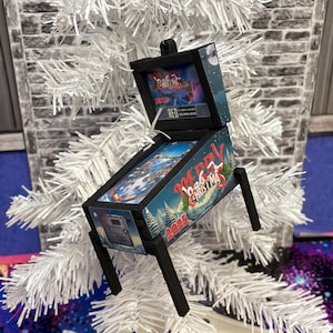 May include: A miniature Christmas-themed pinball machine ornament. The ornament is black with a blue and white winter scene and the words "Merry Christmas 2023". It is hanging on a white Christmas tree.