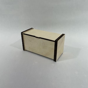 May include: A small, rectangular wooden box with a hinged lid. The box is made of light-colored wood with dark brown trim around the edges. The box is sitting on a white surface.