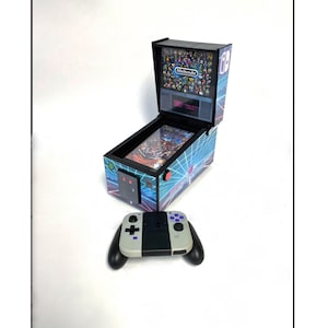 May include: A miniature arcade pinball machine with a Nintendo logo and a screen displaying a grid of characters. The machine is open and the pinball is visible. A Nintendo Switch controller is in the foreground.