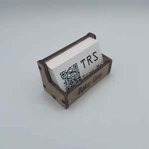 May include: A wooden business card holder with a stack of white cards. The cards have the letters "TRS" and a QR code printed on them. The holder has the words "Take One" printed on the front.