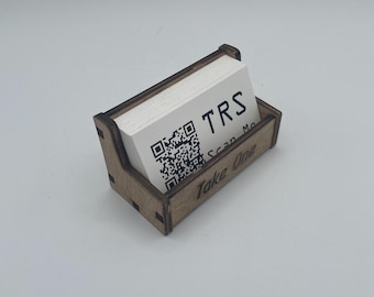 Op de afbeelding: Een houten visitekaarthouder met een stapel witte kaarten. De kaarten hebben de letters "TRS" en een QR-code. De houder heeft de tekst "Take One" op de voorkant.