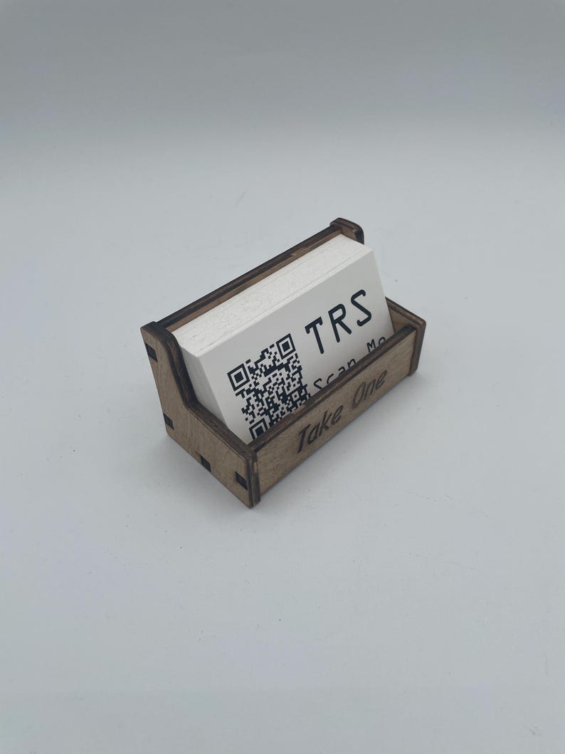 May include: A wooden business card holder with a stack of white cards. The cards have the letters "TRS" and a QR code printed on them. The holder has the words "Take One" printed on the front.