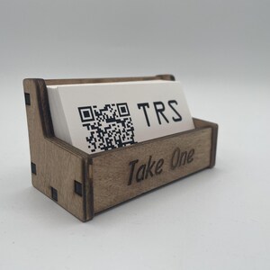 May include: A wooden business card holder with a stack of white cards inside. The cards have a QR code and the letters "TRS" printed on them. The holder is engraved with "Take One".