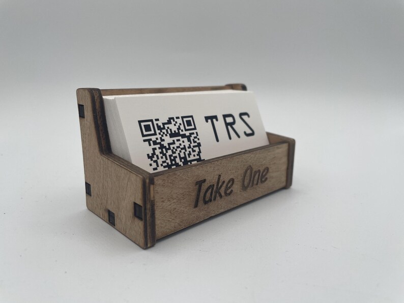 May include: A wooden business card holder with a stack of white cards inside. The cards have a QR code and the letters "TRS" printed on them. The holder is engraved with "Take One".