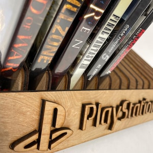 Może przedstawiać: Drewniany uchwyt na gry PlayStation z logo PlayStation i napisem "PlayStation". Zawiera kilka pudełek z grami, w tym "God of War" i "Killzone".