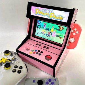 May include: A pink and black arcade cabinet with a screen displaying the game "Super Princess Peach". The cabinet has a joystick and buttons. There are two controllers, one white with yellow buttons and one purple with a black joystick.