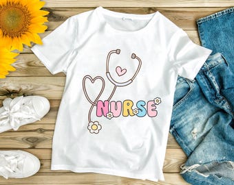 May include: White t-shirt with a colorful graphic of a stethoscope forming a heart shape and the word "NURSE" in pastel colors. The design includes small flower accents. The shirt is displayed on a wooden surface with a sunflower, white sneakers, and blue jeans.
