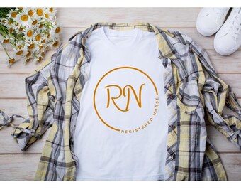 May include: A white V-neck t-shirt with an orange circular design featuring the letters "RN" and the words "Registered Nurse". The shirt is laid on a plaid shirt with white sneakers and a bouquet of daisies.