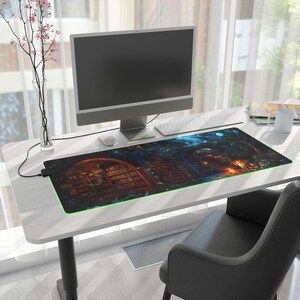 May include: A large, rectangular, black and blue gaming mouse pad with a fantasy scene of a glowing, arched doorway leading to a library with bookshelves. The mouse pad is on a white desk with a computer monitor, keyboard, and a white mug.