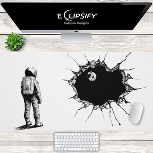 May include: White mouse pad featuring a black and white illustration of an astronaut looking at a hole in space with a moon. The mouse pad has the text "E CLIPSIFY Custom Designs".