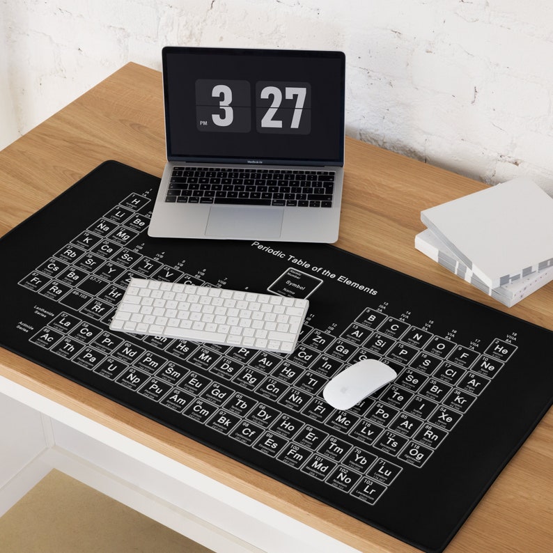 Op de afbeelding: Een zwarte bureau mat met een witte tabel van het periodiek systeem van de elementen erop gedrukt. Een laptop, een wit toetsenbord en een witte muis liggen op de mat. Het laptopscherm toont een digitale klok die 15:27 uur aangeeft.