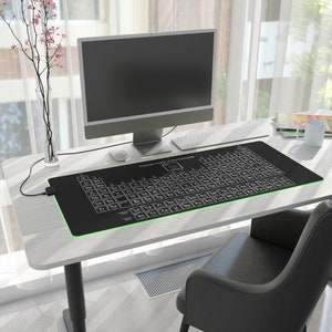 Op de afbeelding: Een zwarte bureaumat met een groene rand heeft een witte tabel van het periodiek systeem van de elementen. De mat ligt op een wit bureau met een computermonitor, toetsenbord en een kopje.