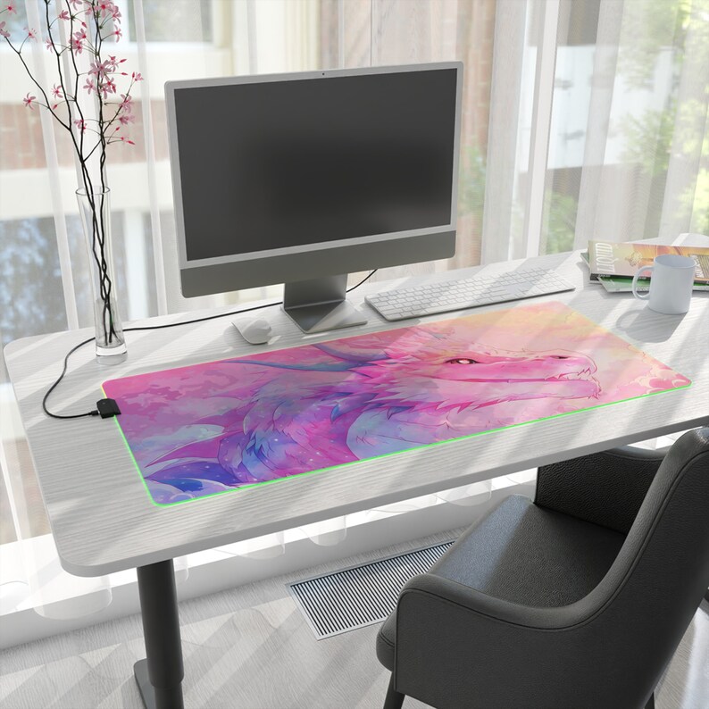 May include: A pink and purple dragon with green eyes is featured on a large rectangular desk mat. The mat has a light pink background with white clouds and a light blue sky. The mat is on a white desk with a computer monitor, keyboard, and a cup on it.