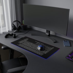 Op de afbeelding: Een zwarte gamingmuis met blauwe accenten ligt op een zwarte muismat met een periodiek systeemontwerp. De muismat is verlicht met blauwe en paarse lampjes. Een zwarte computermonitor en een zwarte mobiele telefoon zijn ook zichtbaar op het bureau.