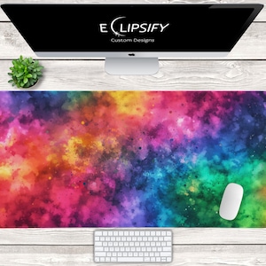 Pode incluir: Um tapete de mesa abstrato colorido em aquarela com um arco-íris de cores, incluindo rosa, amarelo, verde e azul. O tapete tem um design de galáxia com tinta salpicada e um mouse de computador branco no lado direito.