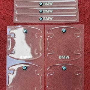Puede incluir: Protectores transparentes y adhesivos para manijas de puertas de automóviles con el logotipo de BMW. El juego incluye cuatro protectores de manijas y cinco protectores largos y delgados, todos con el emblema de BMW en azul, blanco y negro. Sobre fondo rojo.