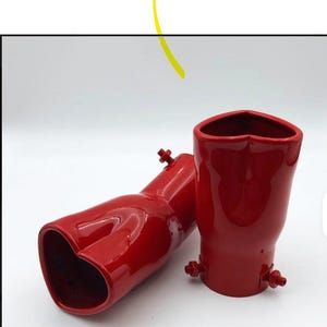 May include: Two red exhaust tips with a heart-shaped opening. The tips are made of a glossy material and have mounting hardware. One tip is lying on its side, while the other stands upright.