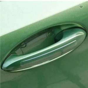 May include: A metallic green car door handle featuring a clear, adhesive protective film applied to the recessed area behind the handle to prevent scratches. The handle is integrated into the smooth, glossy green body panel of the vehicle.