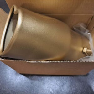 May include: A gold-colored metal object, possibly a light fixture or decorative element, rests inside a cardboard box. The object has a flared, bell-like shape with a rounded top and a threaded fitting. The box is open, revealing the item.