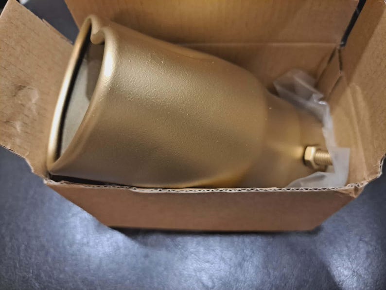 May include: A gold-colored metal object, possibly a light fixture or decorative element, rests inside a cardboard box. The object has a flared, bell-like shape with a rounded top and a threaded fitting. The box is open, revealing the item.