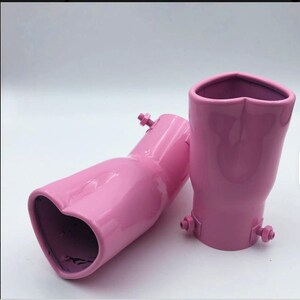 May include: Two pink exhaust tips with heart-shaped openings. The tips are made of metal and have a glossy finish. Each tip has a mounting bracket with a bolt. The background is plain white.