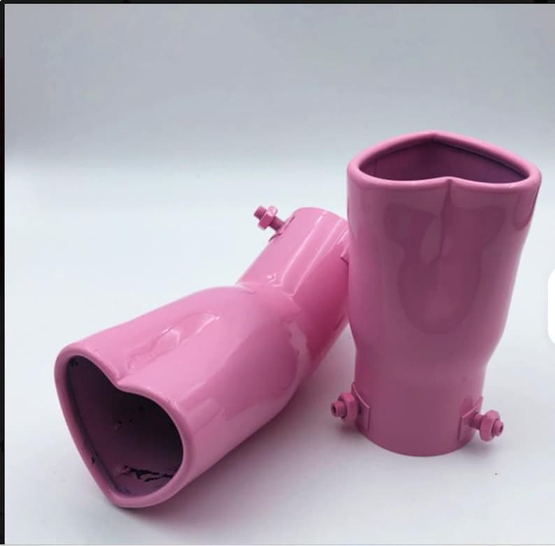 May include: Two pink exhaust tips with heart-shaped openings. The tips are made of metal and have a glossy finish. Each tip has a mounting bracket with a bolt. The background is plain white.