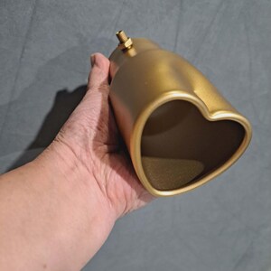 May include: A gold-colored exhaust tip with a heart-shaped opening. The exhaust tip has a cylindrical body and a threaded fitting on top. The object is held in a hand against a gray background.