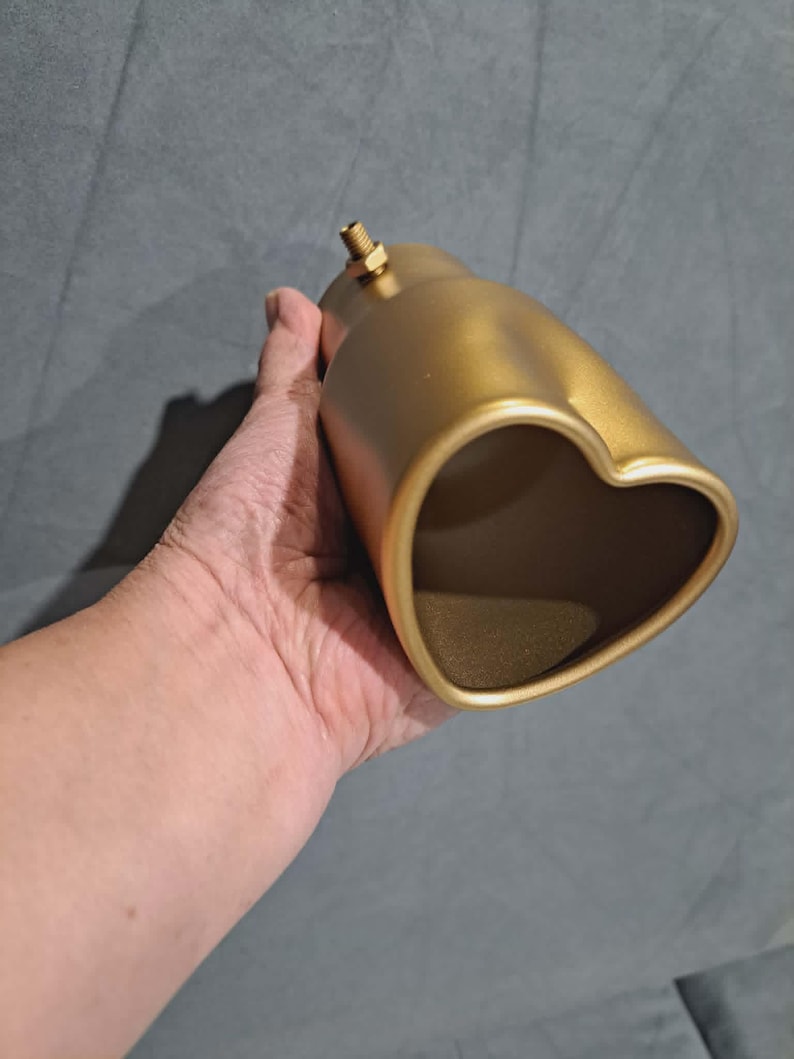 May include: A gold-colored exhaust tip with a heart-shaped opening. The exhaust tip has a cylindrical body and a threaded fitting on top. The object is held in a hand against a gray background.