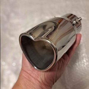 Puede incluir: Una punta de escape cromada pulida con una salida en forma de corazón. El accesorio automotriz es cilíndrico con un extremo acampanado y una abrazadera para la instalación. El metal brillante refleja el entorno.