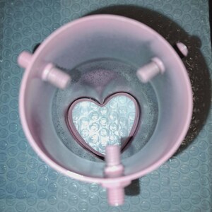 May include: A pink, cylindrical object with three protruding bolt-like structures and a heart-shaped cutout at the base. The object is set against a blue bubble wrap background, creating a contrast in textures and colors.