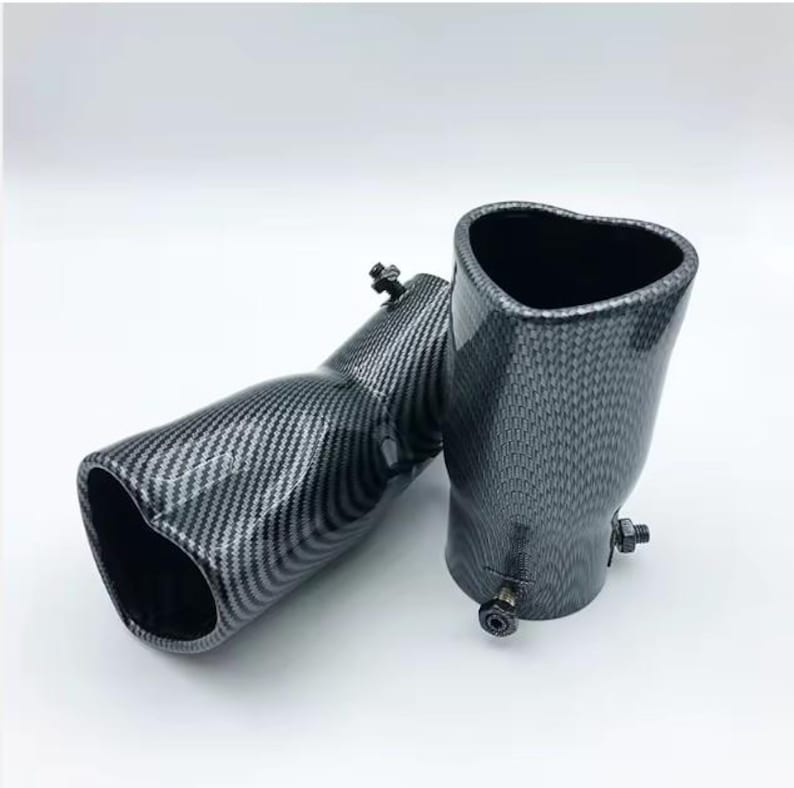 May include: Two carbon fiber exhaust tips with a heart-shaped opening. The tips have a black and gray checkered pattern and are angled. Each tip has a bolt for attachment. The background is white.