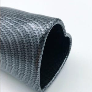 May include: A close-up of a carbon fiber tube with a heart-shaped opening. The tube has a woven pattern in shades of gray and black. The interior of the tube is black.