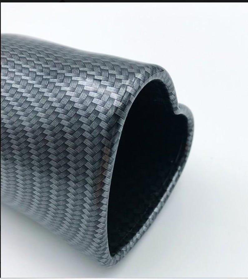 May include: A close-up of a carbon fiber tube with a heart-shaped opening. The tube has a woven pattern in shades of gray and black. The interior of the tube is black.
