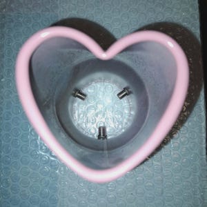 May include: A pink heart-shaped container with three screws visible inside. The container is made of a smooth material and sits on a blue bubble wrap surface. The heart shape is clearly defined with rounded edges.