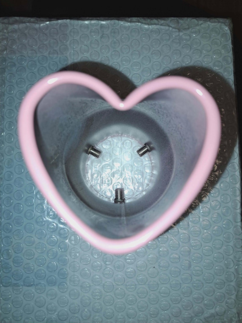 May include: A pink heart-shaped container with three screws visible inside. The container is made of a smooth material and sits on a blue bubble wrap surface. The heart shape is clearly defined with rounded edges.