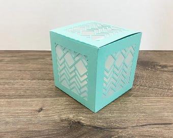 May include: A turquoise paper box with geometric cutouts. The box has a square shape with a patterned design on the sides and top. The box is sitting on a wooden surface.