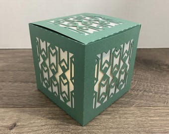 May include: A square, green paper lantern with intricate geometric cutouts. The lantern's design allows light to shine through, creating a decorative effect. The lantern is a cube shape and sits on a wooden surface.
