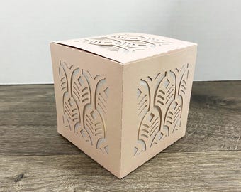 May include: A light pink, square gift box with intricate leaf-shaped cutouts on the sides and top. The box is made of sturdy paper and is designed to hold small items. The box is sitting on a wooden surface.