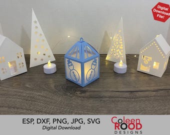May include: A blue paper lantern with a snowman design, surrounded by white paper houses, trees, and candles. The lantern is lit from within, creating a warm glow. The image includes the text "ESP, DXF, PNG, JPG, SVG Digital Download Coleen ROOD DESIGNS".