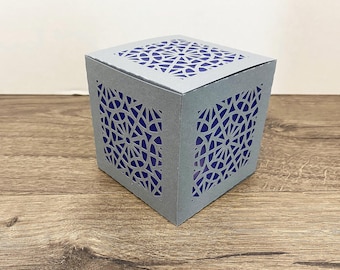 May include: A gray, cube-shaped paper lantern with intricate geometric cutouts. The interior is a deep blue, creating a contrast with the gray exterior. The lantern sits on a wooden surface.
