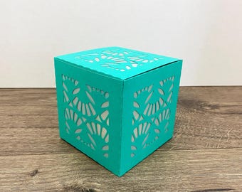May include: A teal-colored, cube-shaped box with intricate cut-out designs. The box is made of paper or cardstock and sits on a wooden surface. The cut-out patterns create a decorative effect, suitable for gifts or small items.