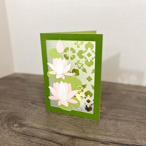 May include: A rectangular greeting card featuring a green frame border. The card displays layered paper cutouts of light pink lotus flowers and green lily pads on the left side, with a decorative green floral pattern on the right side.